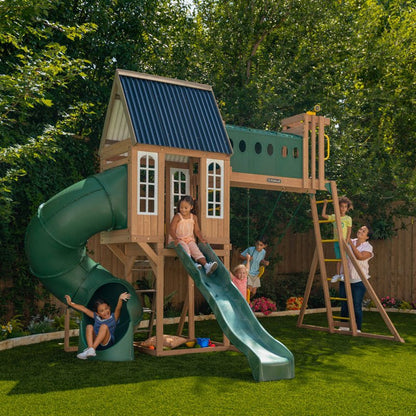 Children play on a wooden playset with a slide, swing, tunnel, and ladder in a grassy backyard.