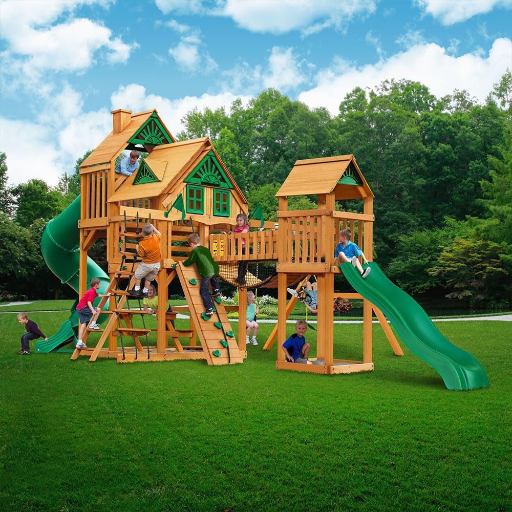 Children play on a wooden playset with green slides, climbing wall, rope ladder, and playhouse features in a grassy yard.