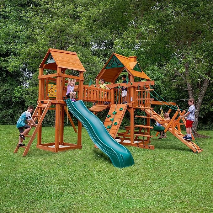Children play on a wooden backyard playset with a slide, climbing wall, ladders, and rope bridge.
