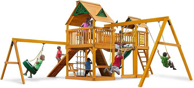 Children play on a wooden playground with swings, a slide, a climbing net, and a playhouse.