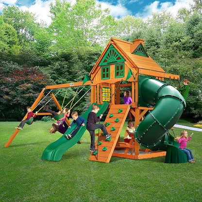 Children playing on a wooden playset with a slide, swing set, climbing wall, and tube slide in a grassy backyard.