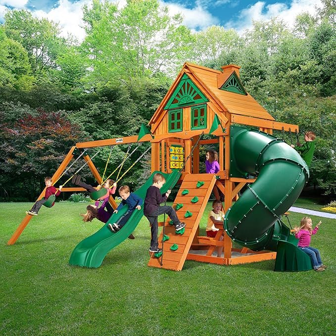Children playing on a wooden playset with a slide, swing set, climbing wall, and tube slide in a grassy backyard.