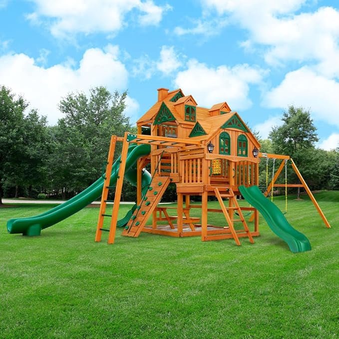 Wooden playset with green slides, climbing wall, and swing set stands on green grass under a blue sky.
