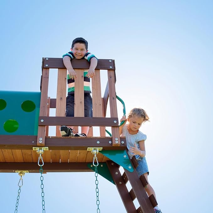 Boy leans over wooden play structure, girl descends ladder against a blue sky.